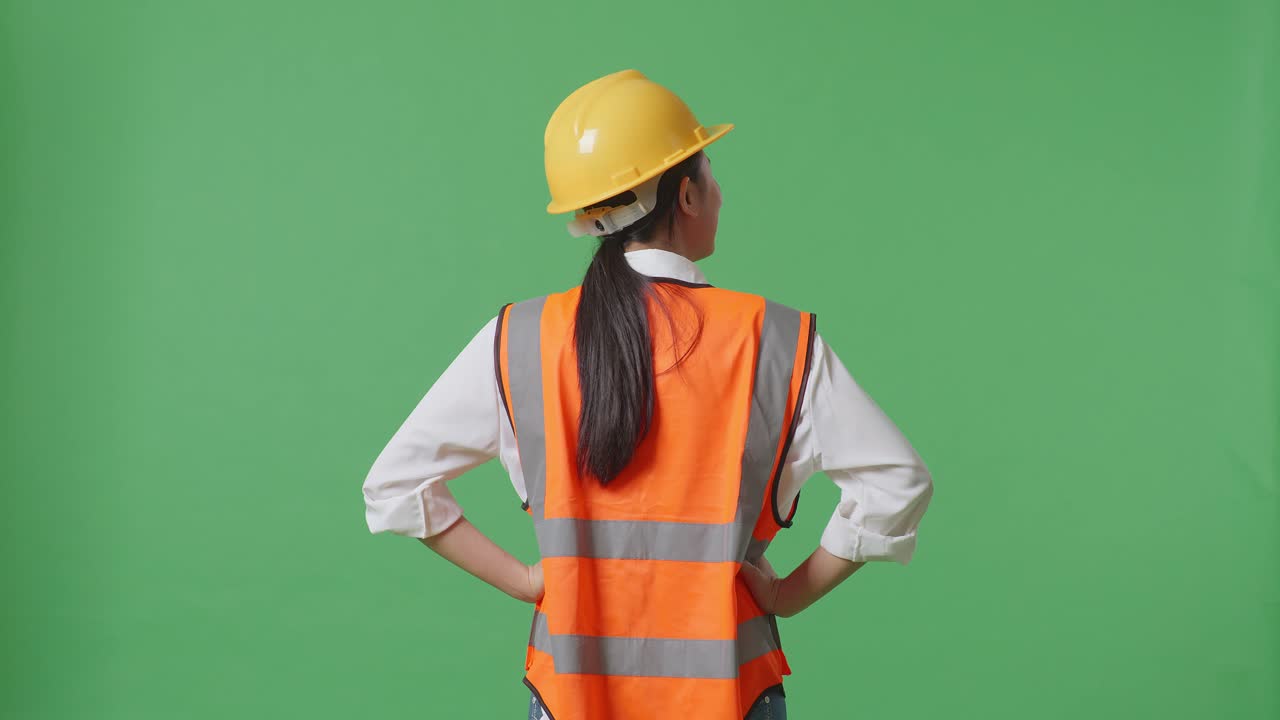 vista trasera de una ingeniera que usa un casco de seguridad mirando a su alrededor mientras está de pie con los brazos akimbo en el estudio de fondo de la pantalla verde