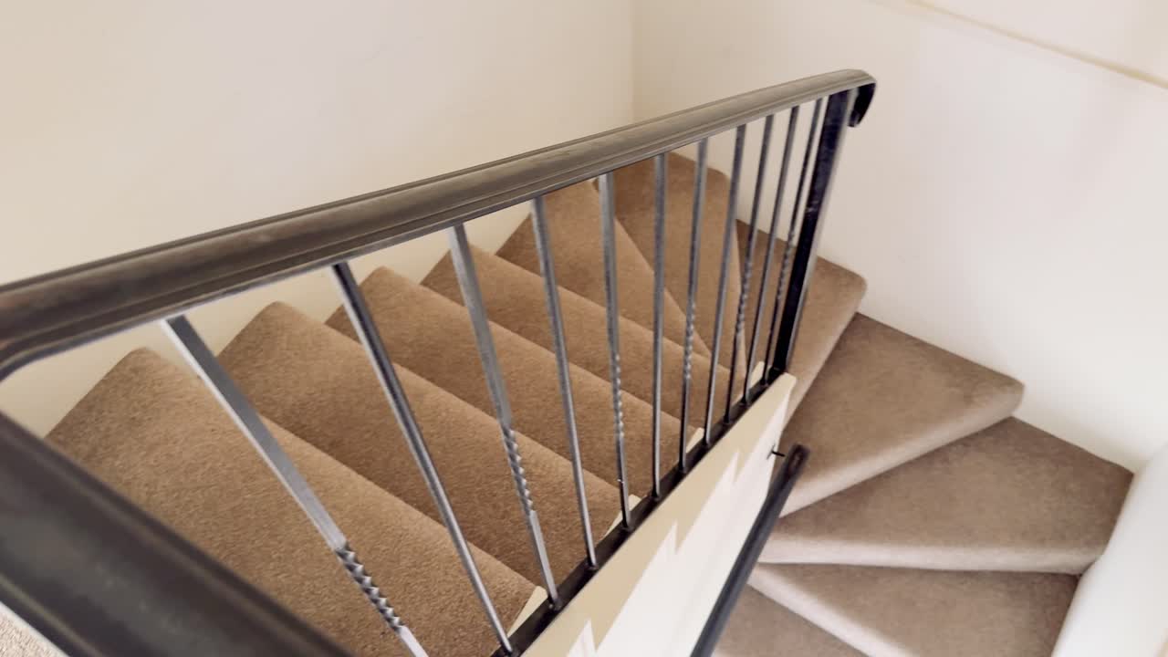 Camera smoothly moves down a modern, carpeted staircase with black metal railing in a well-lit, neutral interior, capturing each step in detail