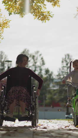 young mommy in wheelchair rests with preschooler daughter in park. woman laughs cheerfully. blonde girl with long braids rides bicycle on warm autumn day