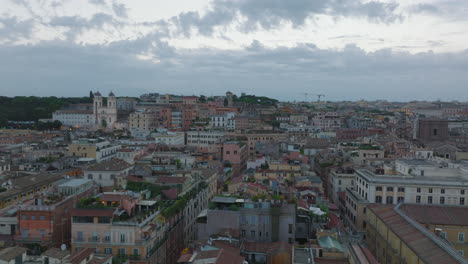 Fly-above-town-development,-old-apartment-buildings-with-rooftop-terraces-and-tourist-landmarks-at-dusk.-Rome,-Italy