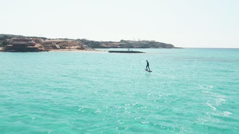 toma aérea de un hombre surfeando en una tabla de surf eléctrica efoil durante un día soleado en cala escondida en ibiza, españa