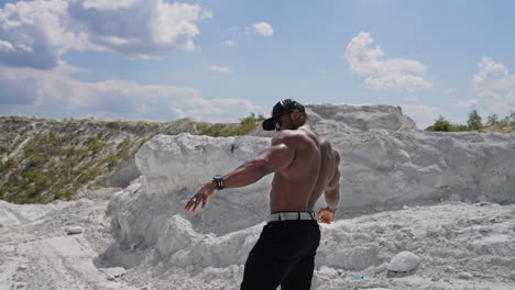 muscular athlete posing on camera. dark skinned sportsman in black cap and sunglasses showing his trained body on white rocky background. circling view.