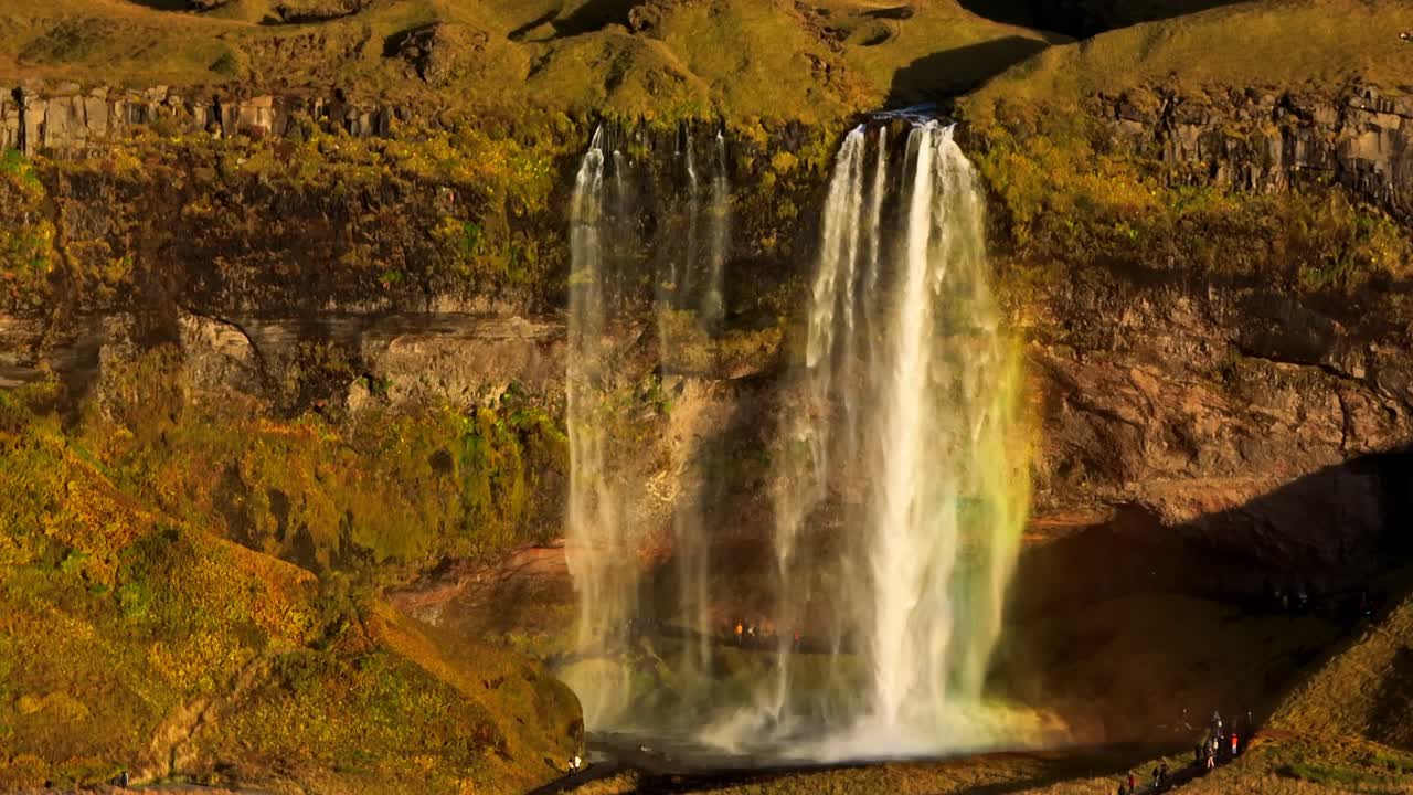 Luchtopname met voorwaartse beweging richting Seljalandsfoss in IJsland, met twee watervalstromen, gloeiende mist en dramatische gouden kliftexturen.