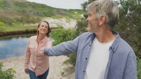smiling senior caucasian couple holding hands and walking at river