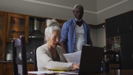 Mixed-race-senior-couple-using-laptop-discussing-and-calculating-finances-at-home