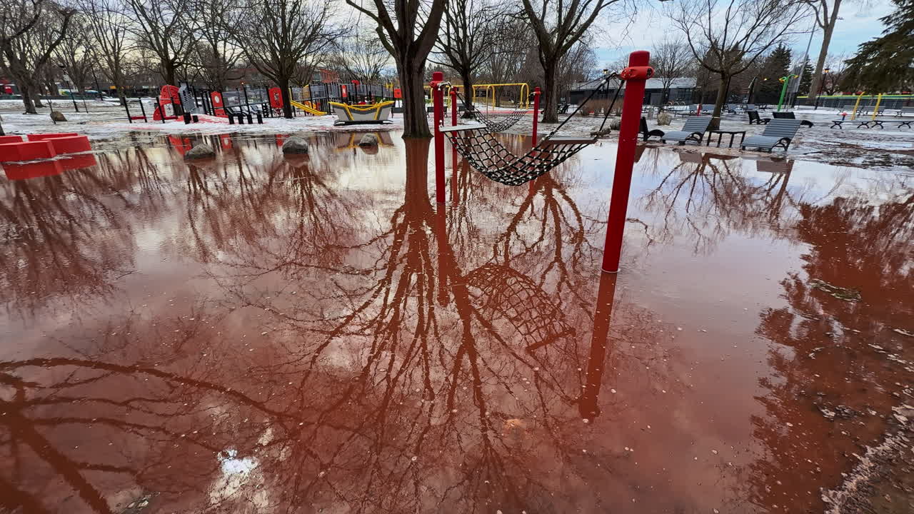Spring arrives all at once, swallowing a public playground in reddish meltwater until the familiar play space turns into a reflective flooded zone no one can use