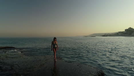 young caucasian woman with long brown hair wearing bathing suit walking on batroun rock outcropping in blue ocean sea water at sunset, lebanon, overhead aerial pull back