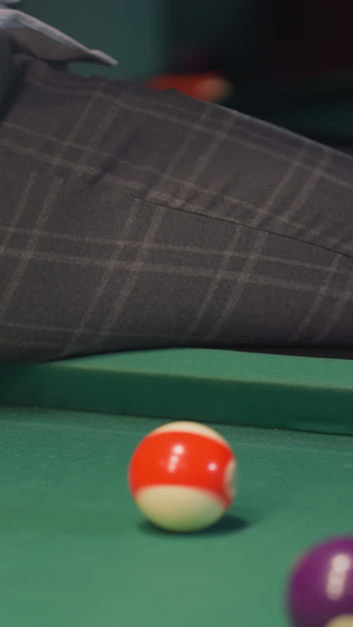 Man in white shirt and grey trousers sits on green pool table, rolling billiard ball. Focus on lower body and hand movement. Stylish indoor setting with soft lighting creating a relaxed ambiance