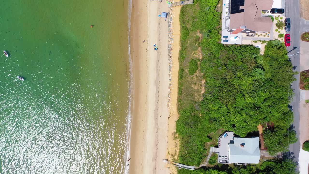 Aerial drone angle shows seaside houses overlooking sandy shoreline, families with umbrellas below, and gentle waves sparkling as they meet the beach