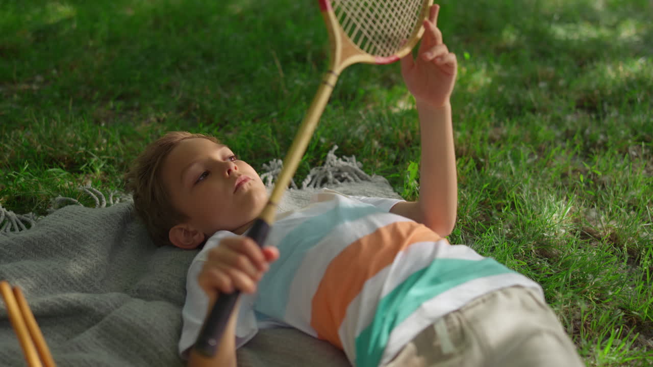 Upset little boy lying blanket in park. Kid examining badminton racket net