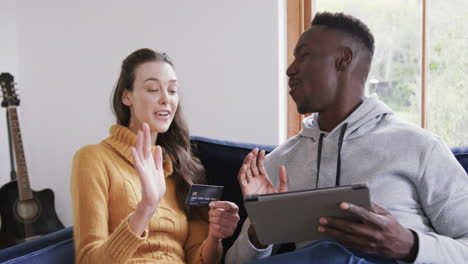 Happy-diverse-couple-sitting-on-sofa,-using-tablet-and-credit-card-in-home,copy-space