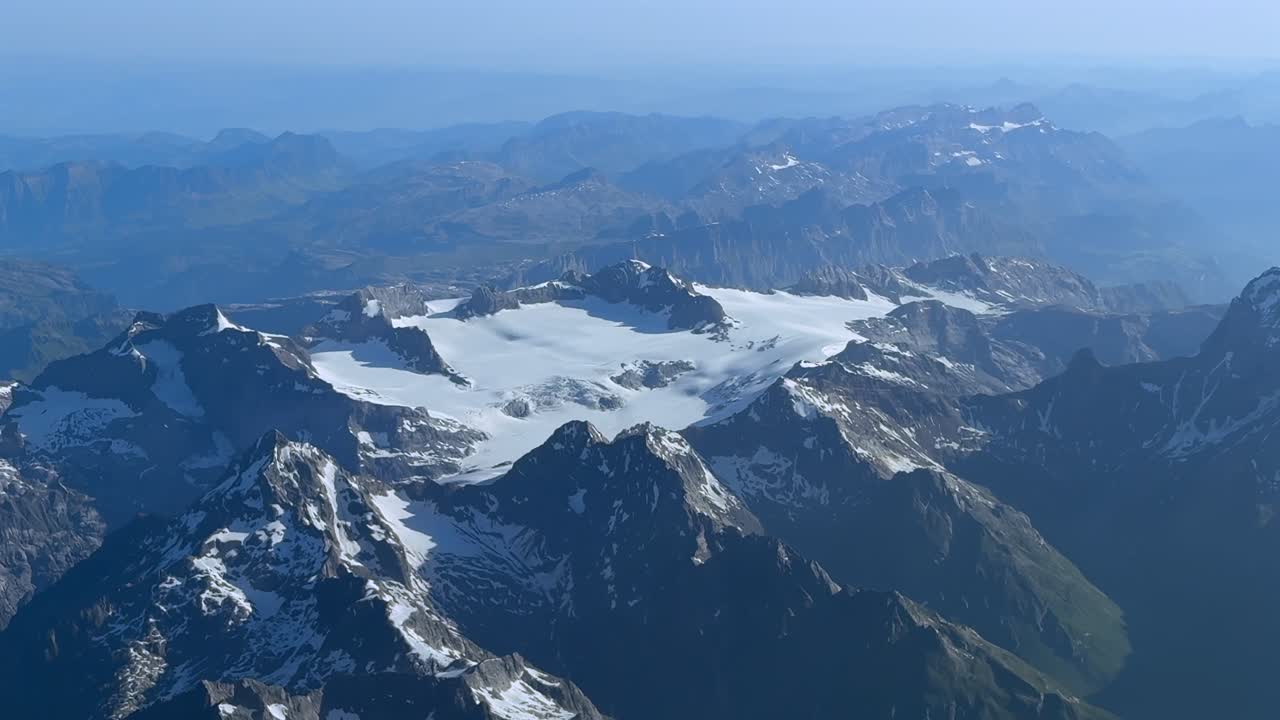 An elevated aerial view of a melting glacier in the northern Swiss Alps in a hazy summer morning. An aerial footage taken from a jet cockpit flying at cruise.