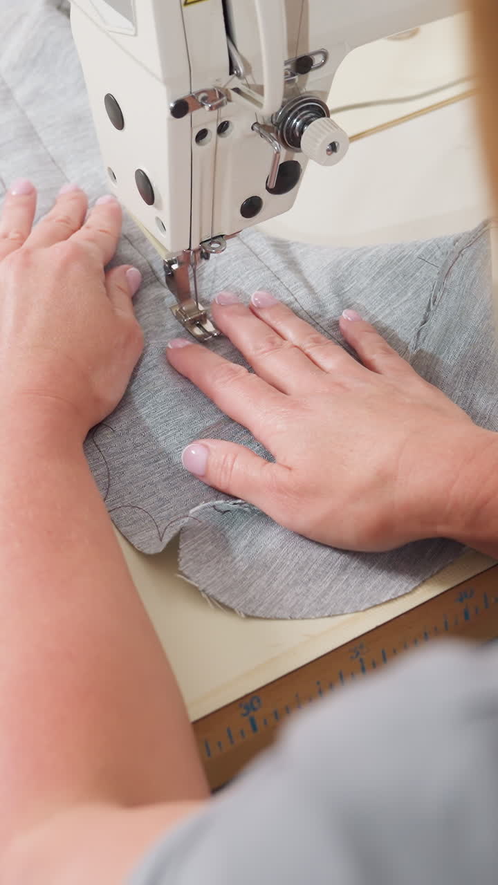 Overhead grey fabric sewing caucasian dressmaker guiding textured ruffle under needle, hands smoothing layered knit while machine feeds material, careful trim and precise stitch shaping couture sample