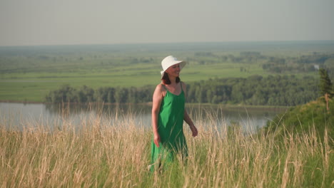 young woman in green gown and white hat walks along grassy cliff edge overlooking wide tranquil river and distant green fields under clear blue sky, appearing thoughtful in scenic landscape