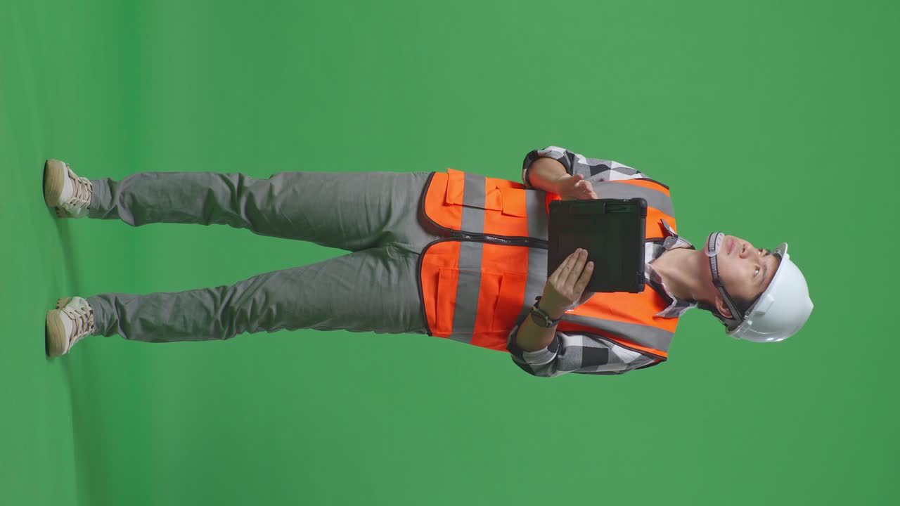 Full Body Of Asian Male Engineer With Safety Helmet Looking At The Tablet In His Hand And Looking Around While Standing In The Green Screen Background Studio