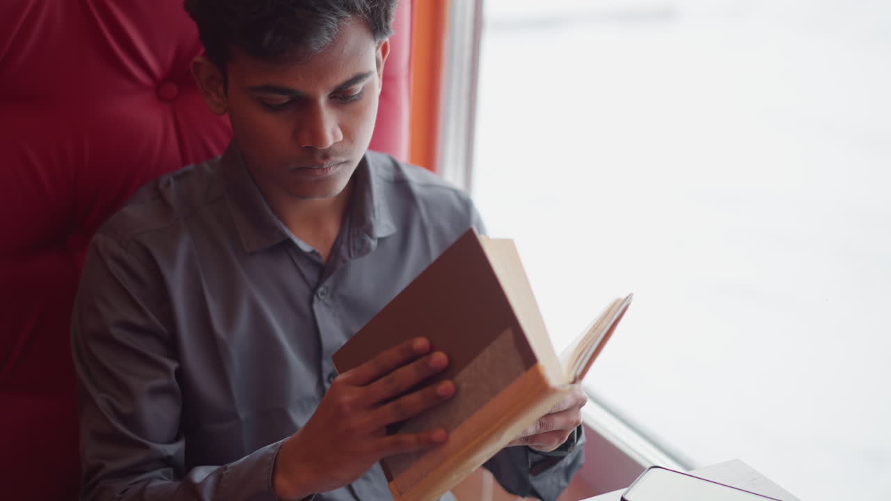 Student sitting in red cushioned booth by bright window during snowy day, deeply focused on reading book, holding pages with care, natural daylight fills quiet space, calm indoor atmosphere, learning moment