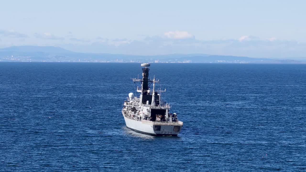 Military vessel sails steadily on blue ocean under clear daylight, distant Scottish coastline visible