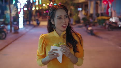 slow motion follow shot of young vietnamese woman in traditional outfit eating local pizza at night in hoi an, vietnam