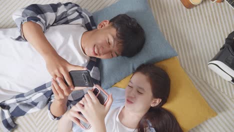 close up top view of asian teen couple playing smartphone while lying on carpet on the floor at home. phone addicted couple