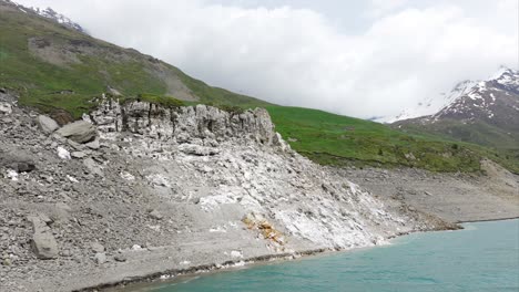 Aerial-View-of-rocky-shoreline-stretching-beside-a-tranquil-lake,-with-lush-green-hills-and-snow-capped-mountains-standing-majestically-under-a-partly-cloudy-sky-in-the-background