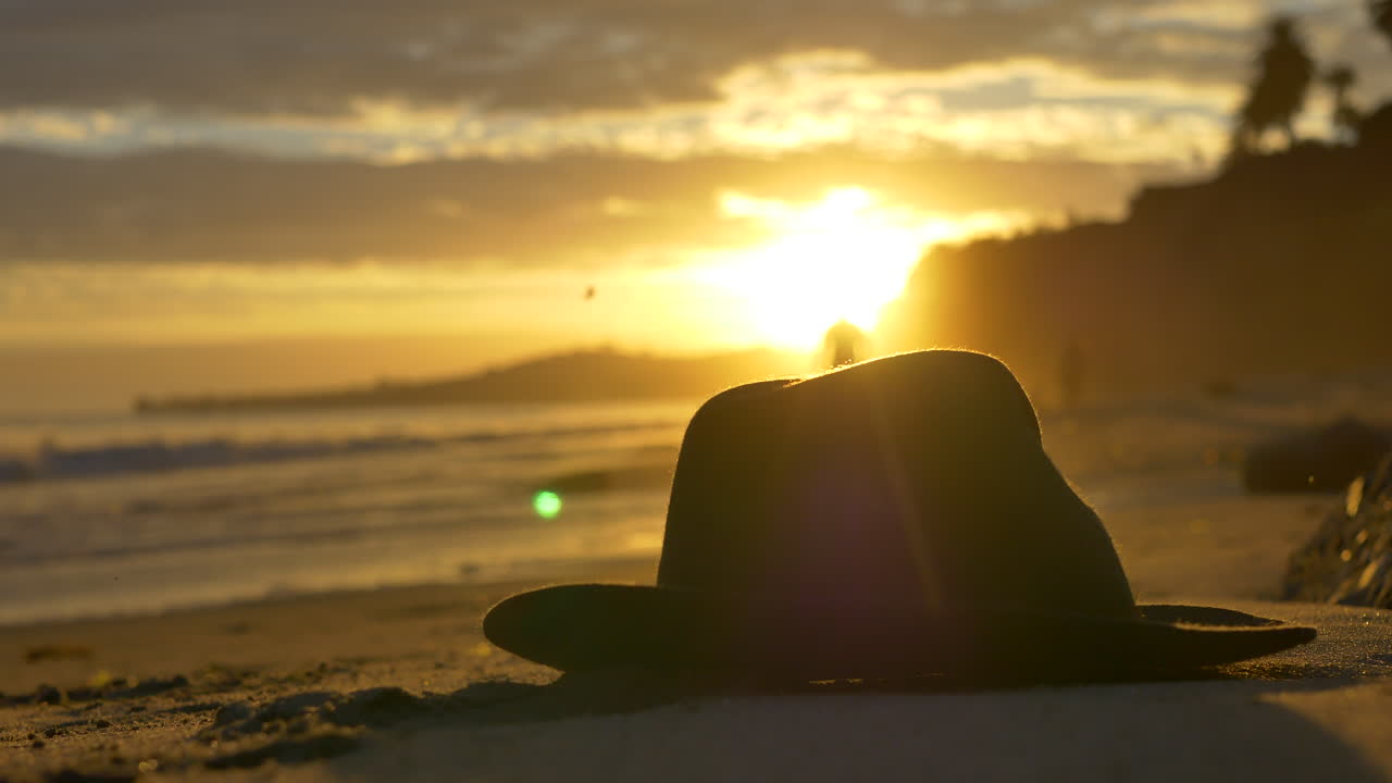 Golden sunset on Butterfly Beach in Montecito, California with a hat in the sand, peaceful ocean waves washing against the shore and people in silhouette walking in the distance