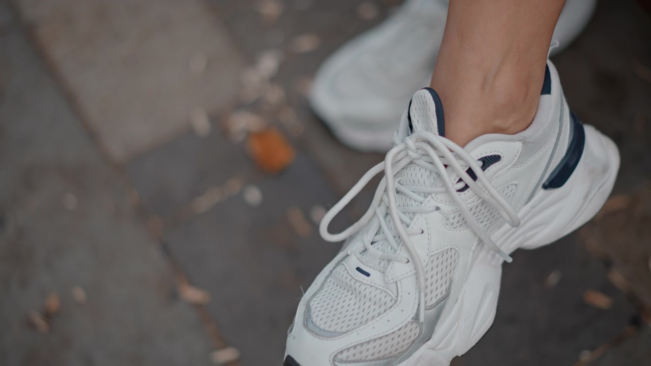 Primo piano ravvicinato dal basso che mostra una mano che tira i lacci per stringere una scarpa da ginnastica bianca, mentre il piede rimane sollevato su un marciapiede urbano esterno con luce naturale e texture dettagliata, con scarpa da ginnastica, scarpa, laccio.