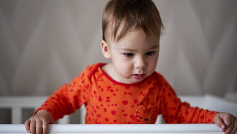 sweet cute boy in orange shirt standing in the cot holding by the handrail. lovely child laughs showing his two lower teeth.
