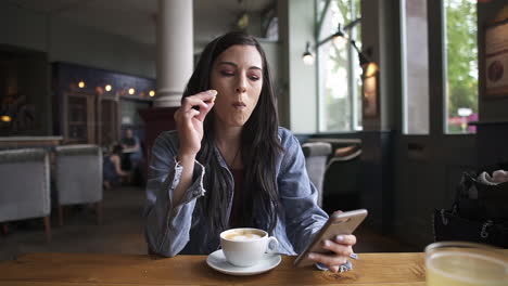 attractive hispanic brunette sitting at the table, eating a cookie and having a coffee while checking her phone