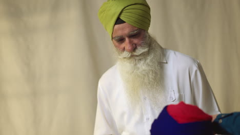 Fotografía-De-Estudio-De-Un-Hombre-Sikh-Mayor-Que-Ayuda-A-Un-Hombre-Sikh-Más-Joven-A-Atar-La-Tela-Para-Un-Turbante-Contra-Un-Fondo-Liso,-Filmada-En-Tiempo-Real-3