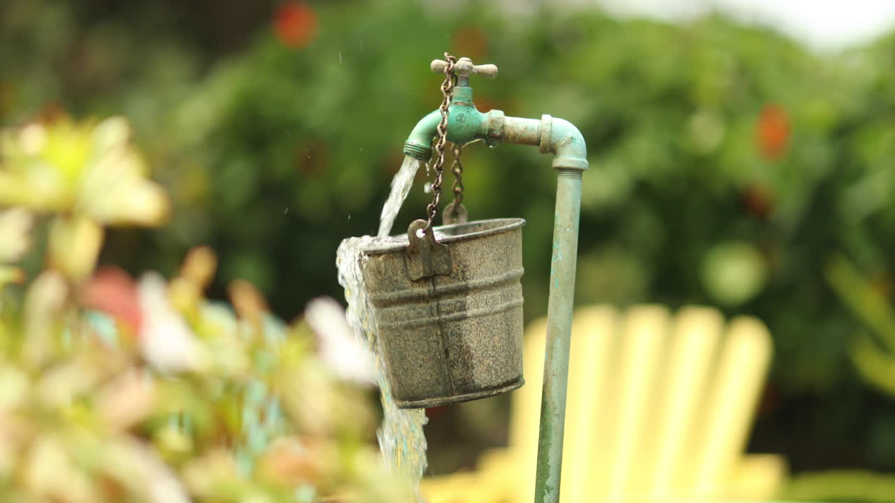 Acqua da un rubinetto da giardino che si riversa e trabocca da un secchio appeso in una botte più grande con sedie a sdraio colorate sullo sfondo.