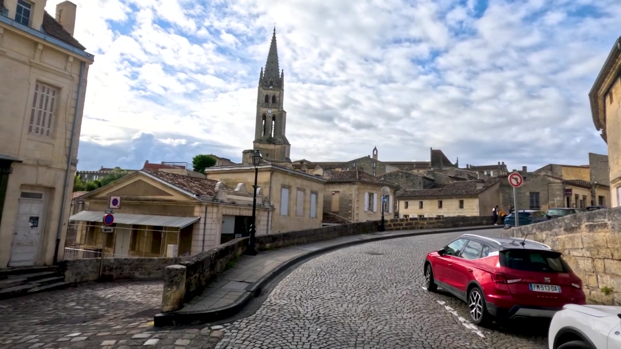 La telecamera si muove lungo una strada acciottolata curva verso il monumentale campanile della chiesa di Saint-Emilion