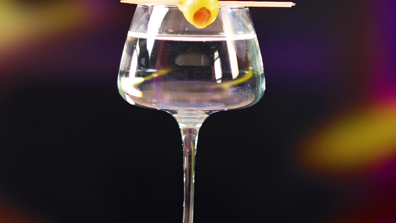 A hand places an olive on a martini glass against a blurred colorful background, highlighting cocktail preparation