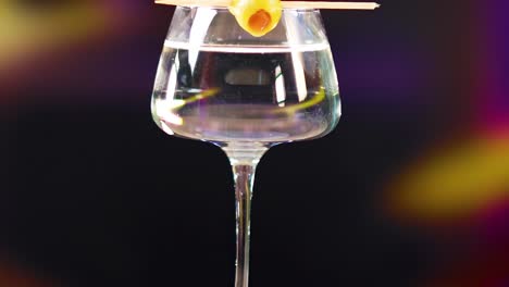 a hand places an olive on a martini glass against a blurred colorful background, highlighting cocktail preparation