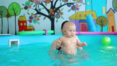 little blond baby boy standing still as coach lifts him up in the swimming pool. trainer teaching a child to swim.