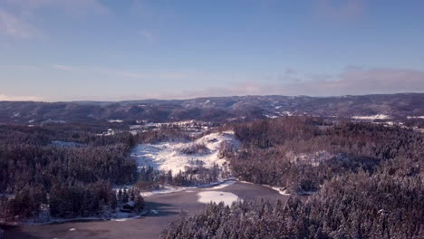 Aerial,-drone-shot-overlooking-snow,-forest,-hills-and-a-pond,-sunny-day,-in-Risvann,-Birkeland,-Agder,-South-Norway