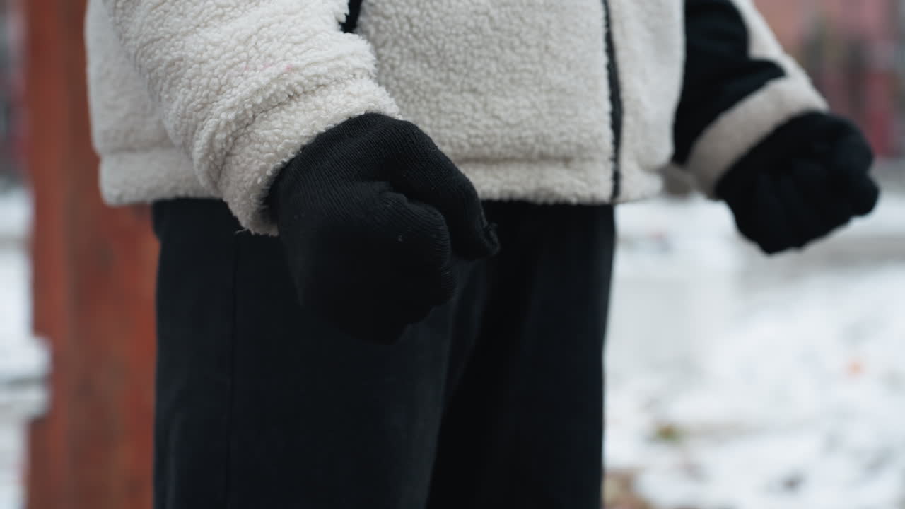 검은 장갑, 겨울 재, 검은 바지를 입은 사람이 손목을 회전시키고, 흐릿한 눈 바탕에 멀리 있는 인물과 지나가는 차를 보여주는 근접적인 측면 시각