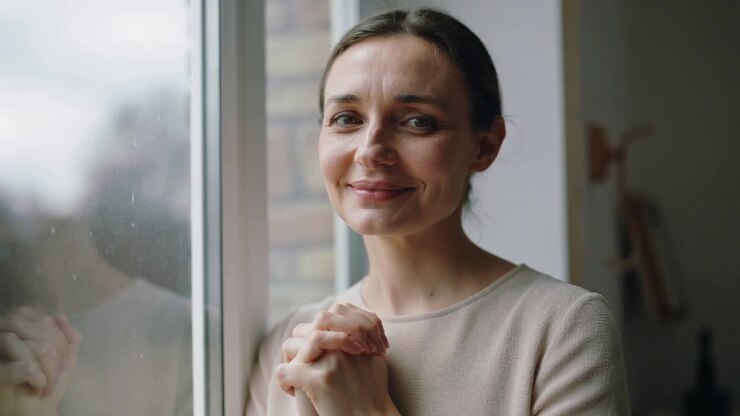 Mujer de mediana edad sonriente junto a la ventana, notando su reflejo, juntando las manos, pensativa con un suéter beige.