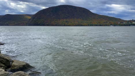 Rocky-river-banks-on-a-windy-day-with-choppy-water-and-hills,-mountains,-and-a-village-on-the-other-side