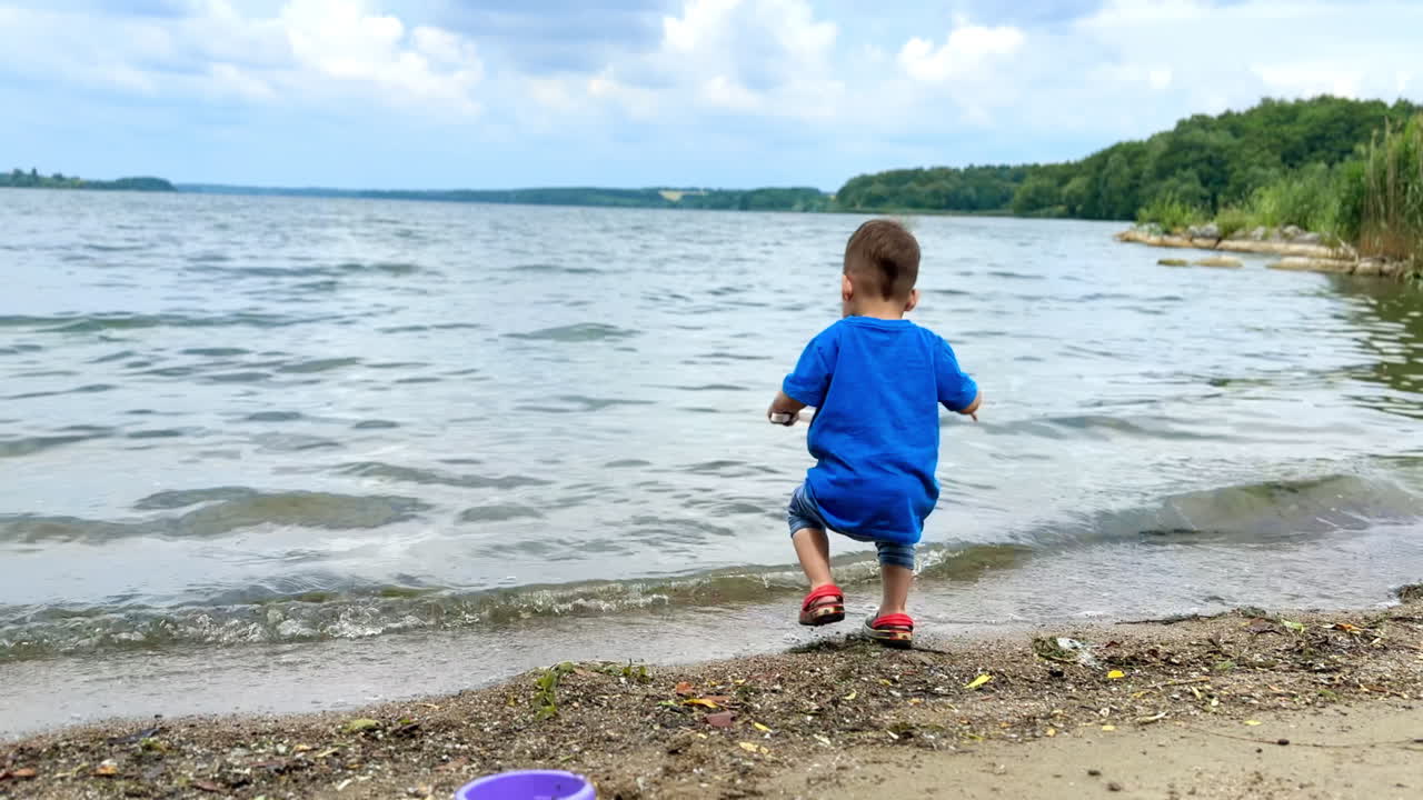 Little toddler in blue t-shirt plays at the river bank. Child takes his toy shovel and walks to the water.