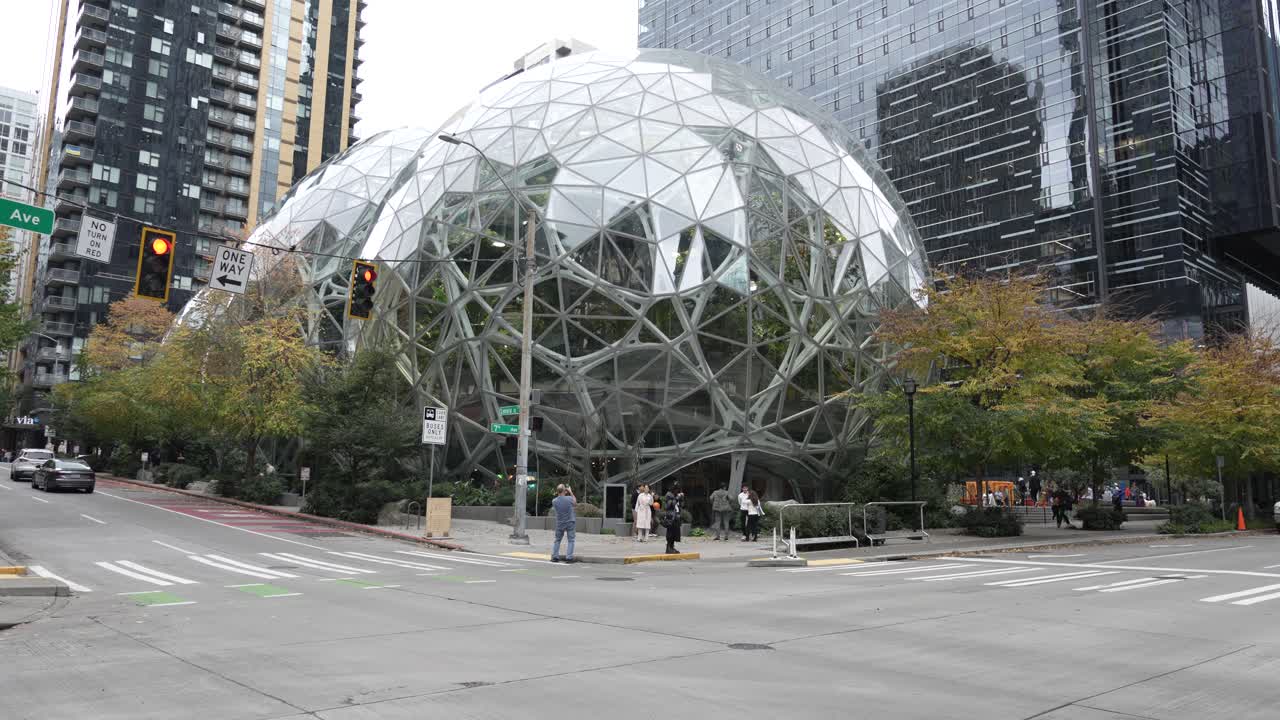 The Spheres at Amazon in Seattle, featuring pedestrians and cars along Lenora St and 7th Ave. The modern glass domes sit amid tall skyscrapers, blending nature with urban architecture.