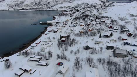 Drone-view-in-Tromso-area-in-winter-flying-over-a-snowy-village-with-wooden-houses-and-the-ocean-on-the-side-in-Norway