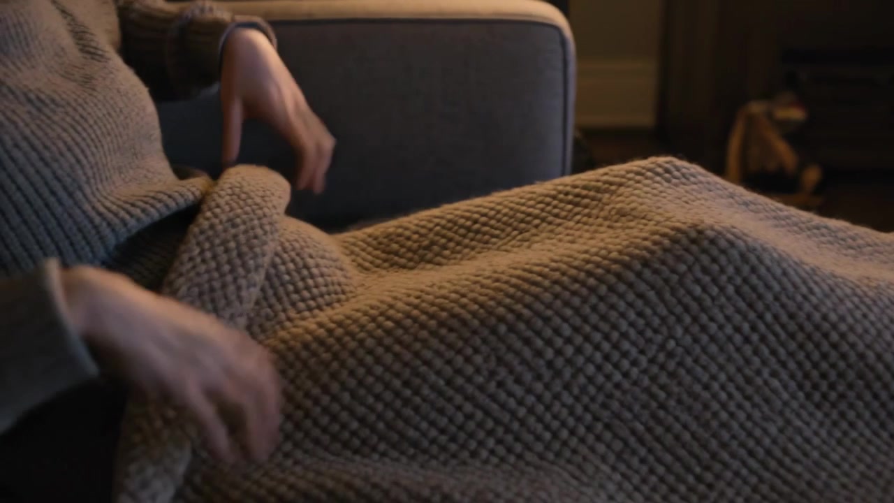 Donna in maglione con orologio che si avvicina, tirando una coperta di maglia sulle gambe, sistemandosi sul divano per il calore.