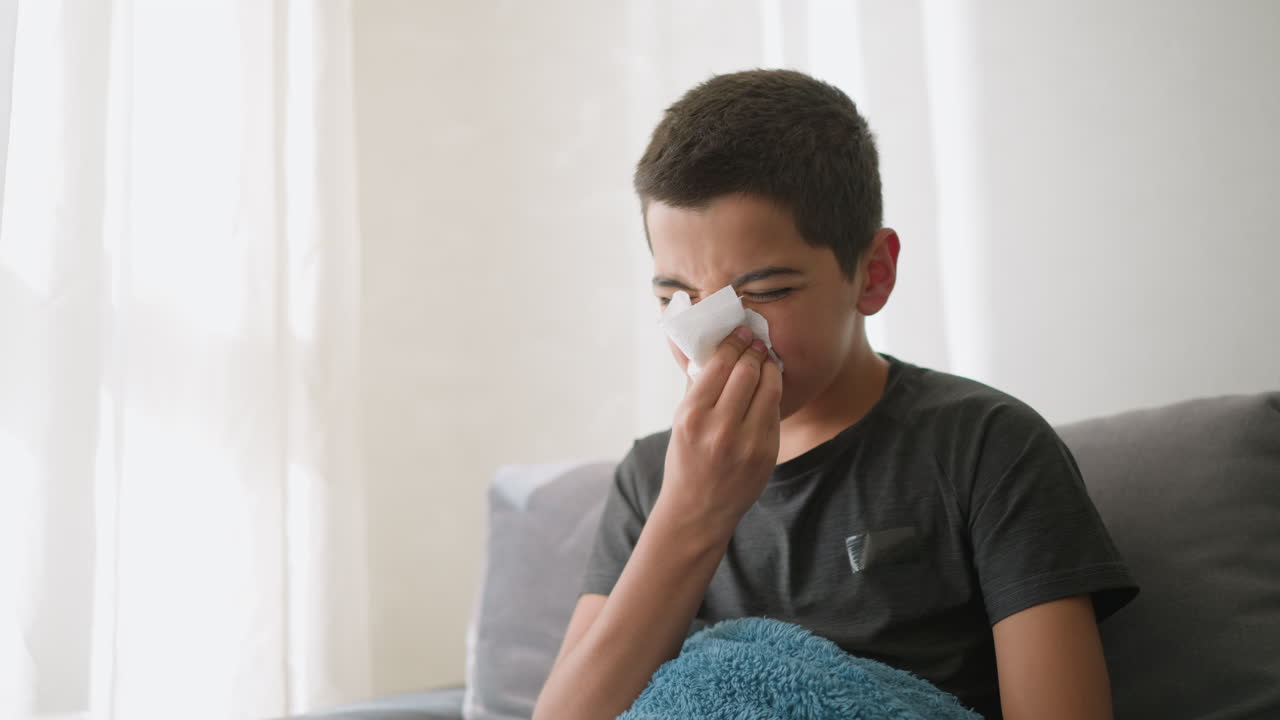 Sick young boy sneezing into tissue, covered with blanket on couch, looks weak and tired, using napkin to wipe nose, sitting by the window in a cozy home setting