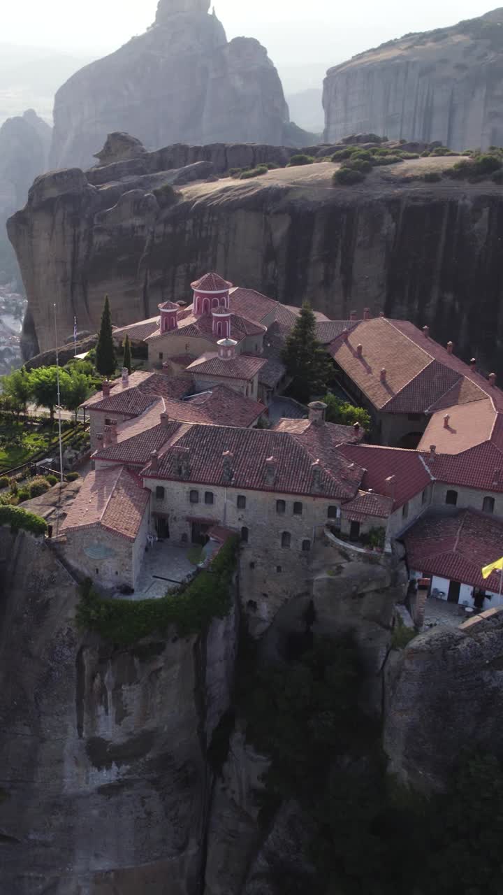 Vertical aerial reveal of Agios Stefanos Monastery on a precipice above Kalabaka, capturing the rocky terrain and town scenery