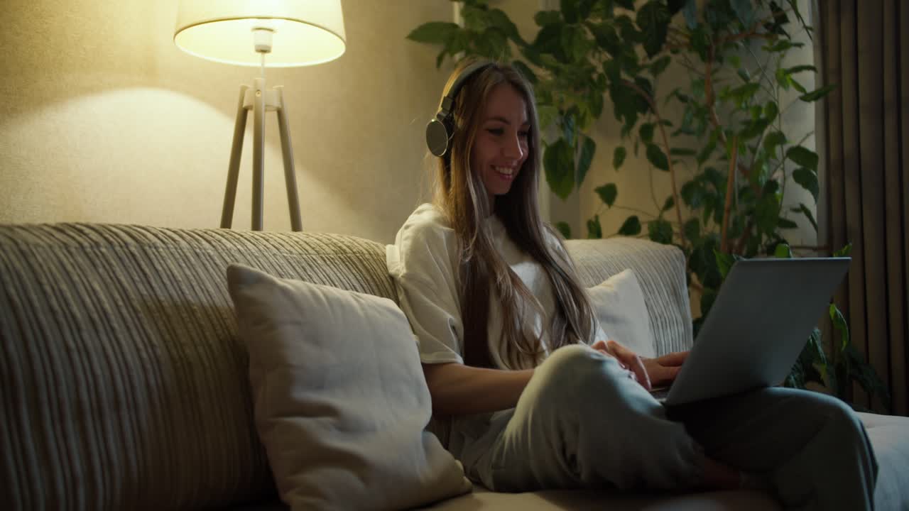 Bela mulher de meia-idade sentada no sofá em casa, ouvindo música com fones de ouvido sem fio e digitando no laptop