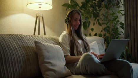 hermosa mujer de mediana edad sentada en el sofá en casa, escuchando música con auriculares inalámbricos y escribiendo en una computadora portátil