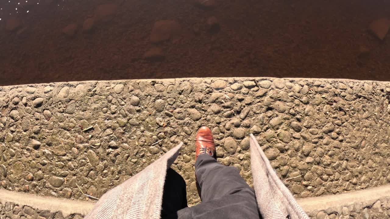 Top-down view of brown leather shoes standing at the edge of a rocky riverside, overlooking shallow brown water under sunlight.