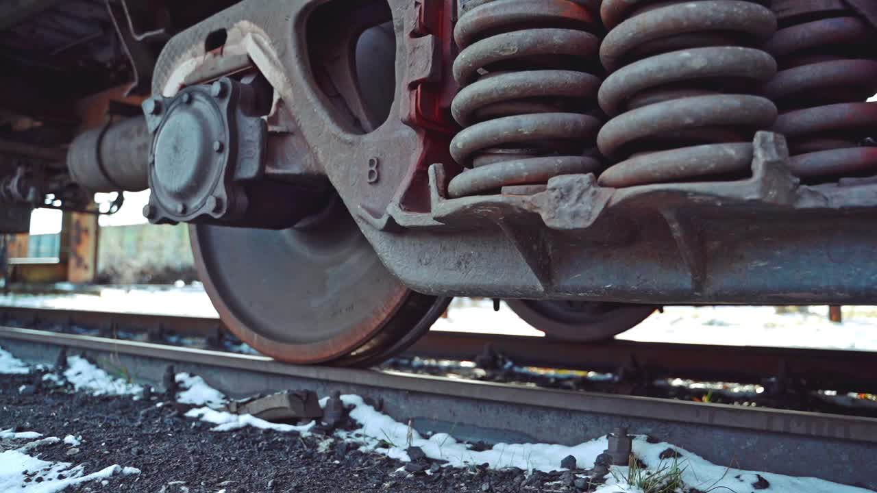 View of bottom to movement of powerful wheel mechanism of a freight car on the background of the ground with snow on the rail station. Close-up.