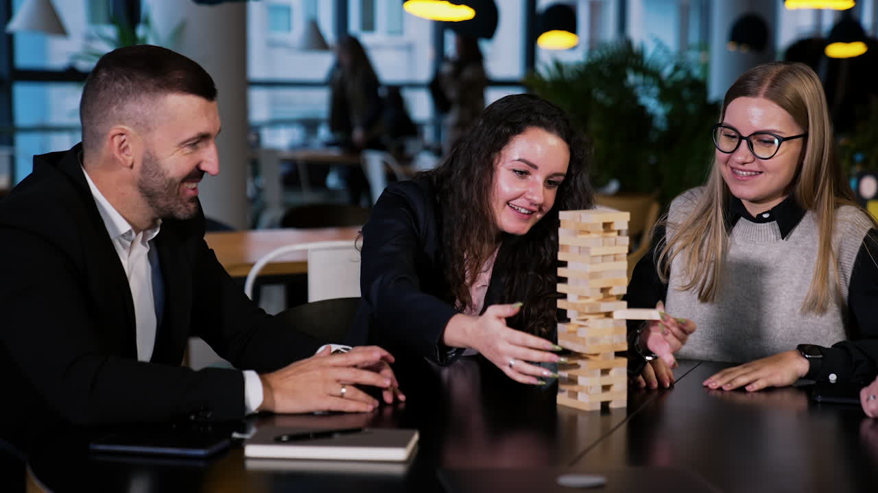 Jenga game played by adult group of people. Dark-haired woman pulls the little brick carefully to place it on top. Other players laugh and have good time.