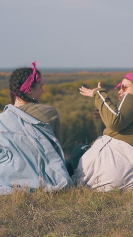 young lesbian exhilarated couple sits on river bank against pictorial landscape under clear sky in autumn backside view